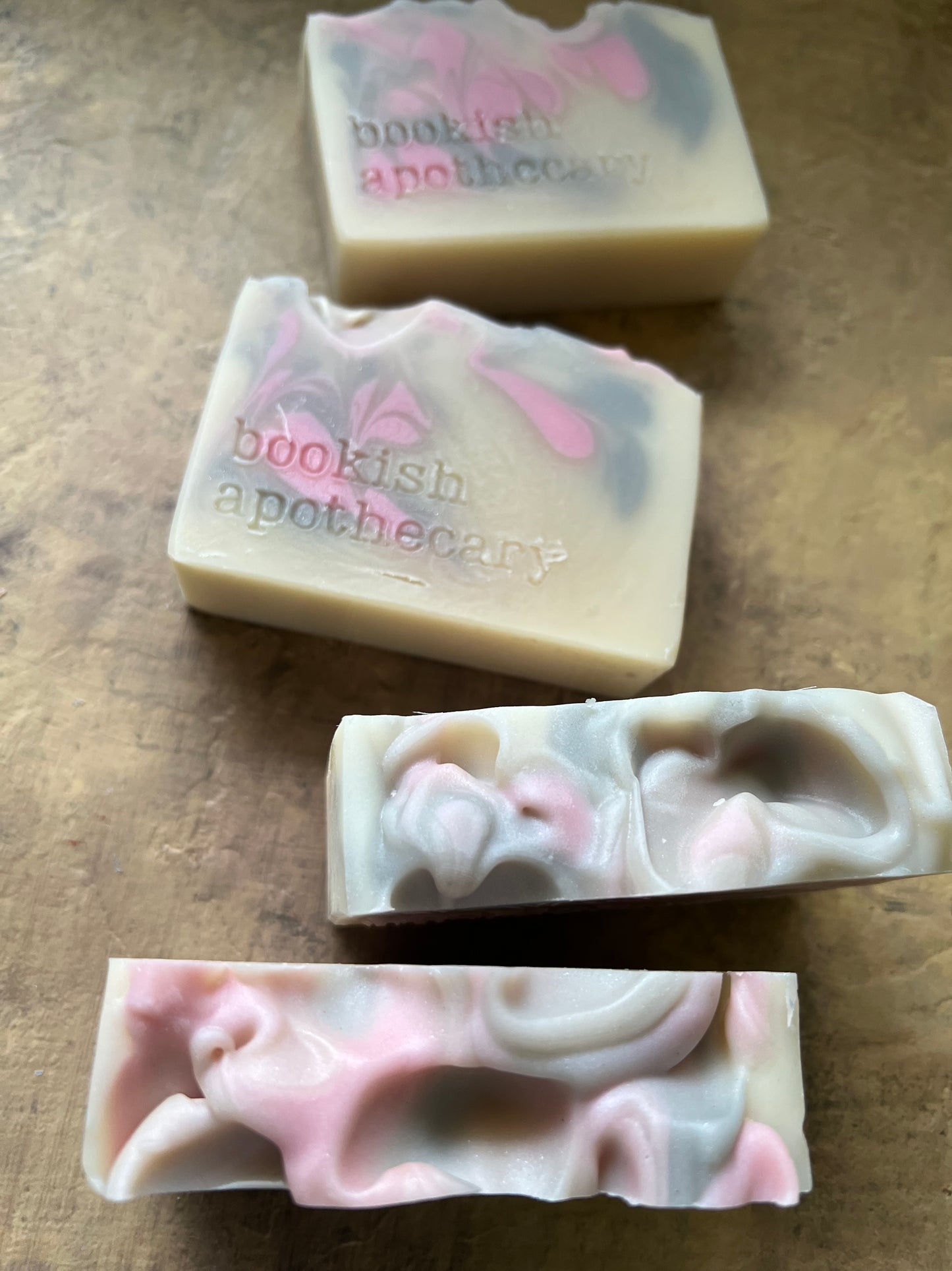 Four handmade soap bars with pink, gray, and off-white marbled swirls, featuring the stamped text 'bookish apothecary' on two of the bars, arranged on a textured brown surface.