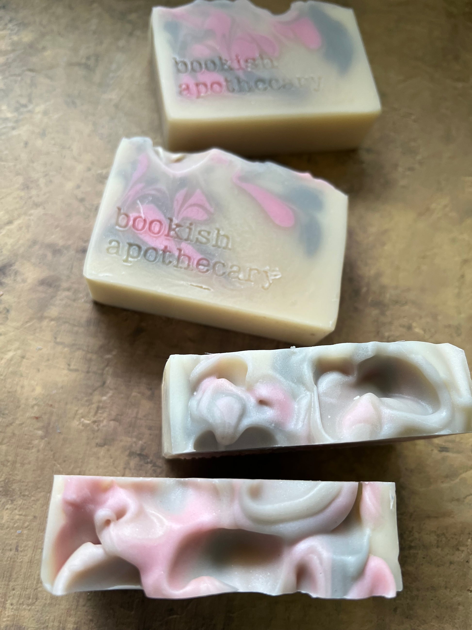 Four handmade soap bars with pink, gray, and off-white marbled swirls, featuring the stamped text 'bookish apothecary' on two of the bars, arranged on a textured brown surface.