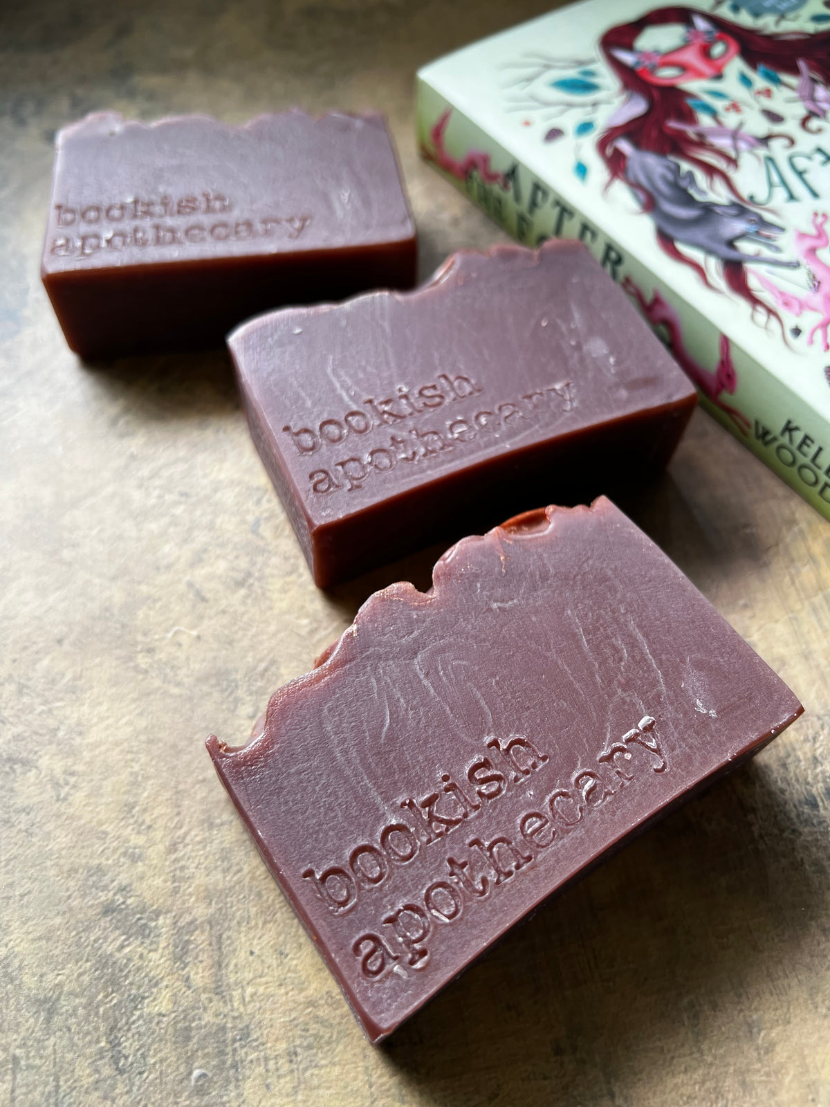Three rectangular bars of dark brown handmade soap , each embossed with the brand name 'bookish apothecary', arranged on a textured brown surface next to a decorative book.