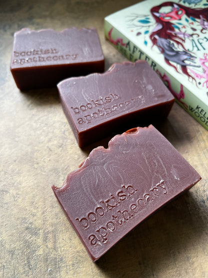 Three rectangular bars of dark brown handmade soap , each embossed with the brand name 'bookish apothecary', arranged on a textured brown surface next to a decorative book.
