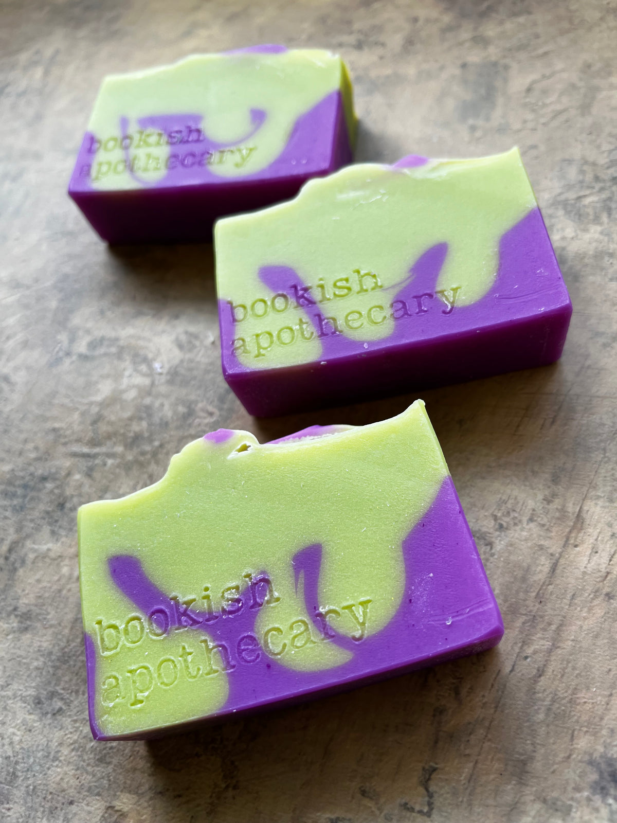Three handmade soap bars with a marbled purple and light green pattern, embossed with the brand name 'bookish apothecary', arranged diagonally on a textured brown surface.