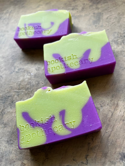 Three handmade soap bars with a marbled purple and light green pattern, embossed with the brand name 'bookish apothecary', arranged diagonally on a textured brown surface.