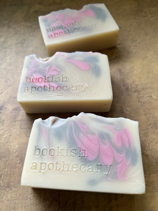 Three handmade artisanal soap bars with swirled pink, gray, and off-white patterns, featuring the brand name 'bookish apothecary' stamped on the side, arranged on a textured brown surface.