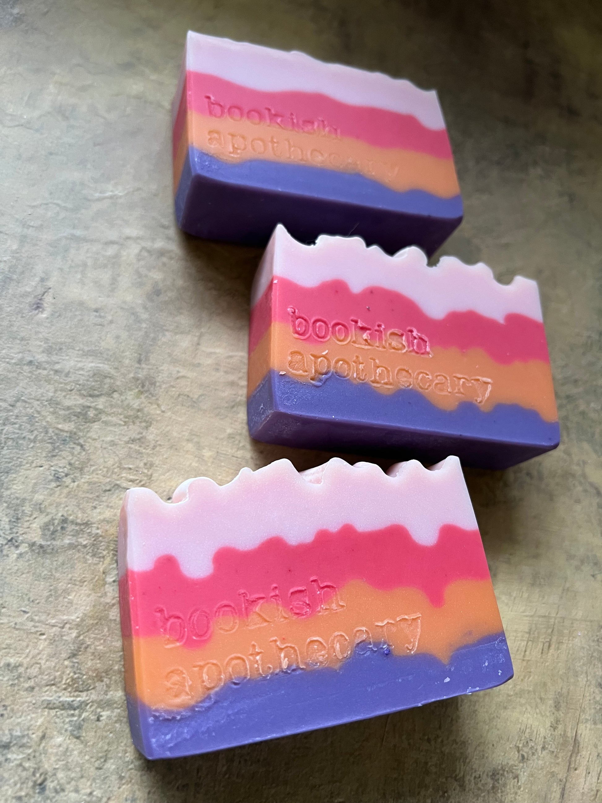 Three handmade soap bars with a wavy color-blocked pattern of purple, pink, orange, and white, resting on a textured gray surface. The brand name 'bookish apothecary' stamped on the sides