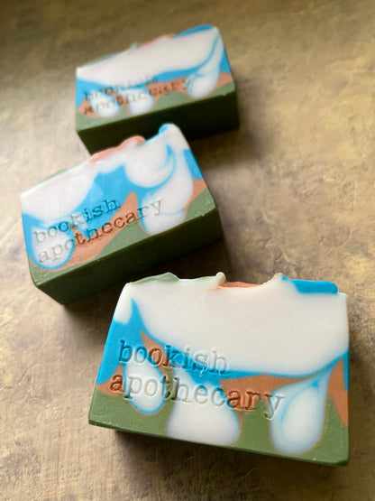 Three handmade soap bars with a swirled pattern of white, light blue, sage green, and peach, featuring the text 'bookish apothecary' stamped on the side, arranged on a textured beige surface.