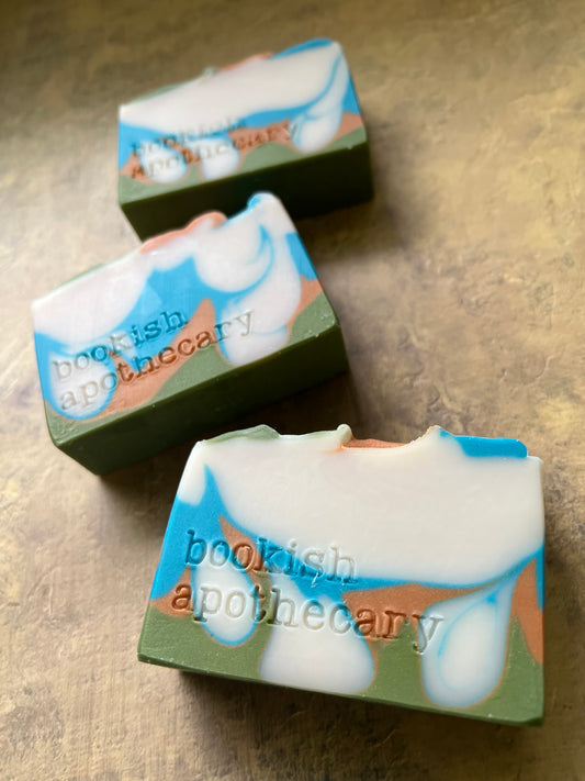 Three handmade soap bars with a swirled pattern of white, light blue, sage green, and peach, featuring the text 'bookish apothecary' stamped on the side, arranged on a textured beige surface.