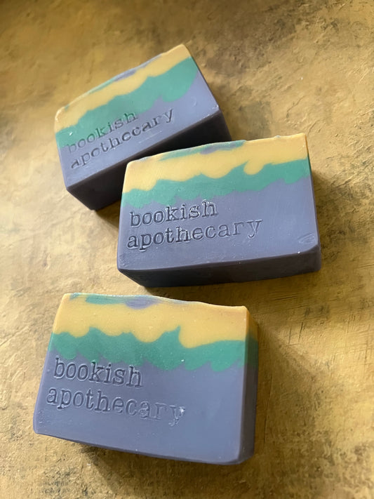Three handmade soap bars with a lavender-purple base and wavy stripes of yellow and teal, embossed with the brand name 'bookish apothecary', arranged on a textured gold surface.