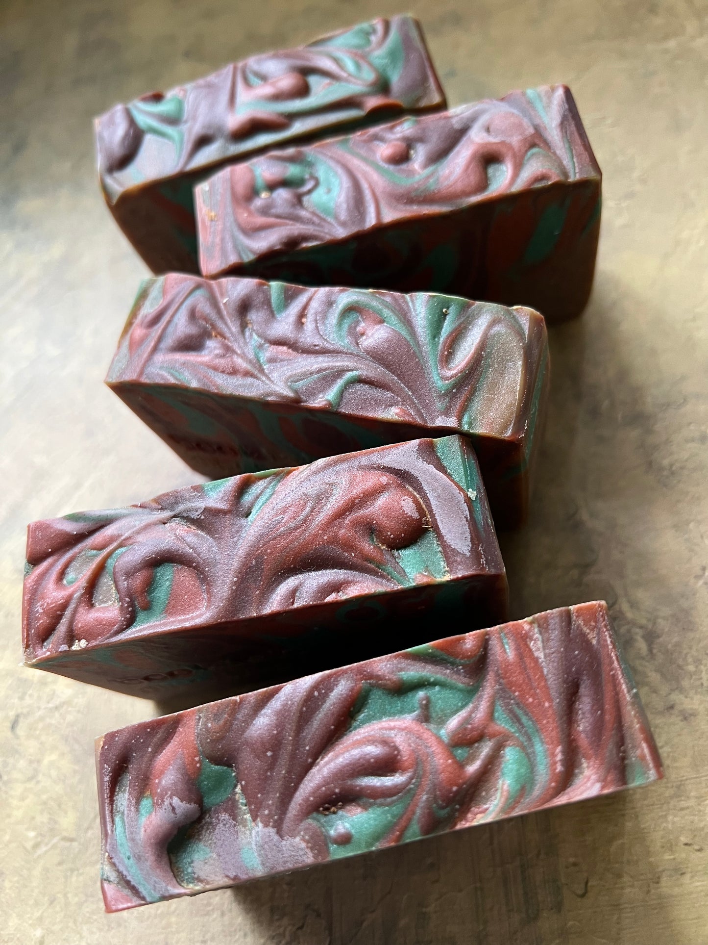 Five rectangular handmade soap bars with a marbled swirl pattern of reddish-brown, teal green, and white, arranged diagonally on a textured gray surface.