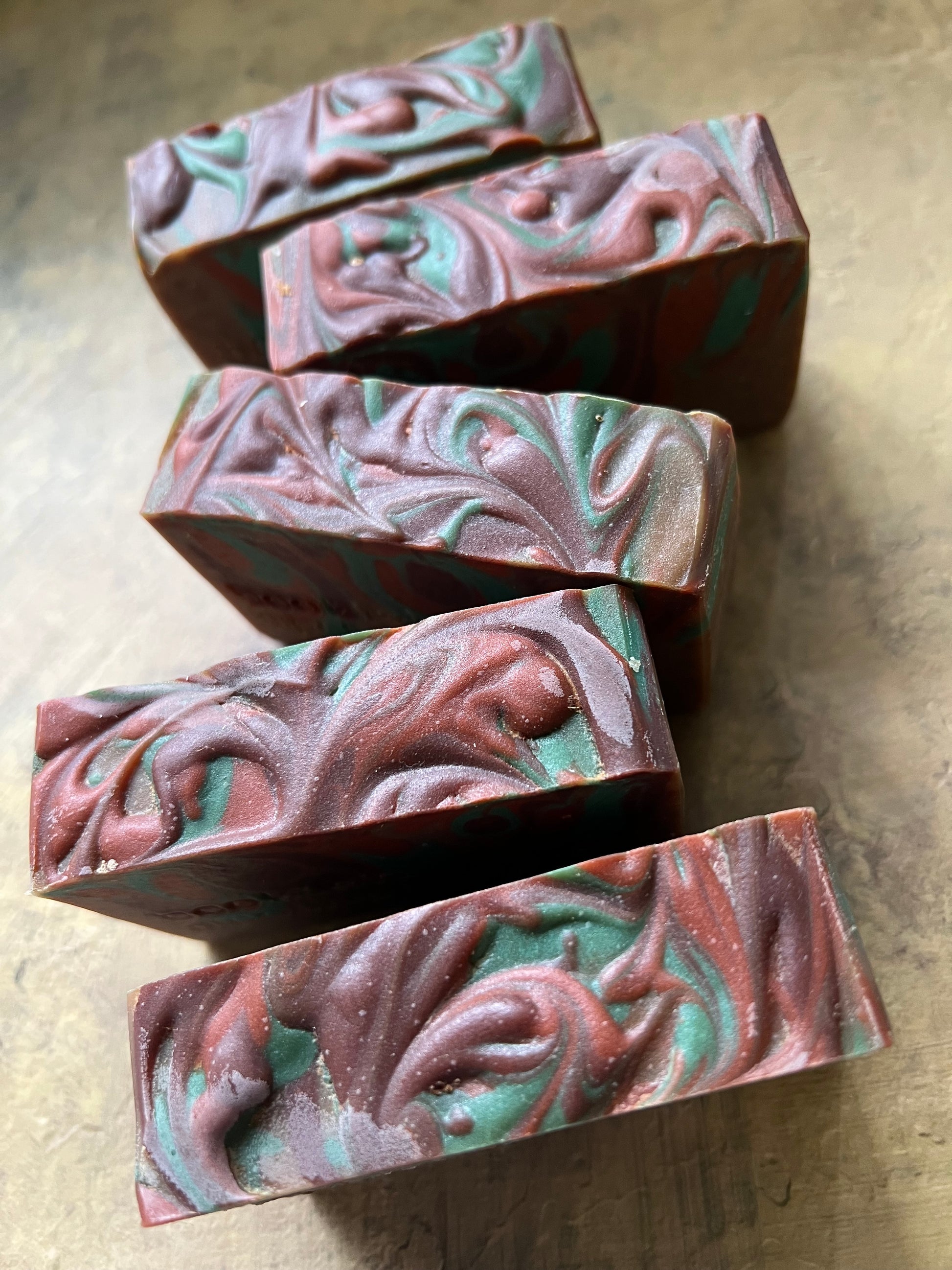 Five rectangular handmade soap bars with a marbled swirl pattern of reddish-brown, teal green, and white, arranged diagonally on a textured gray surface.