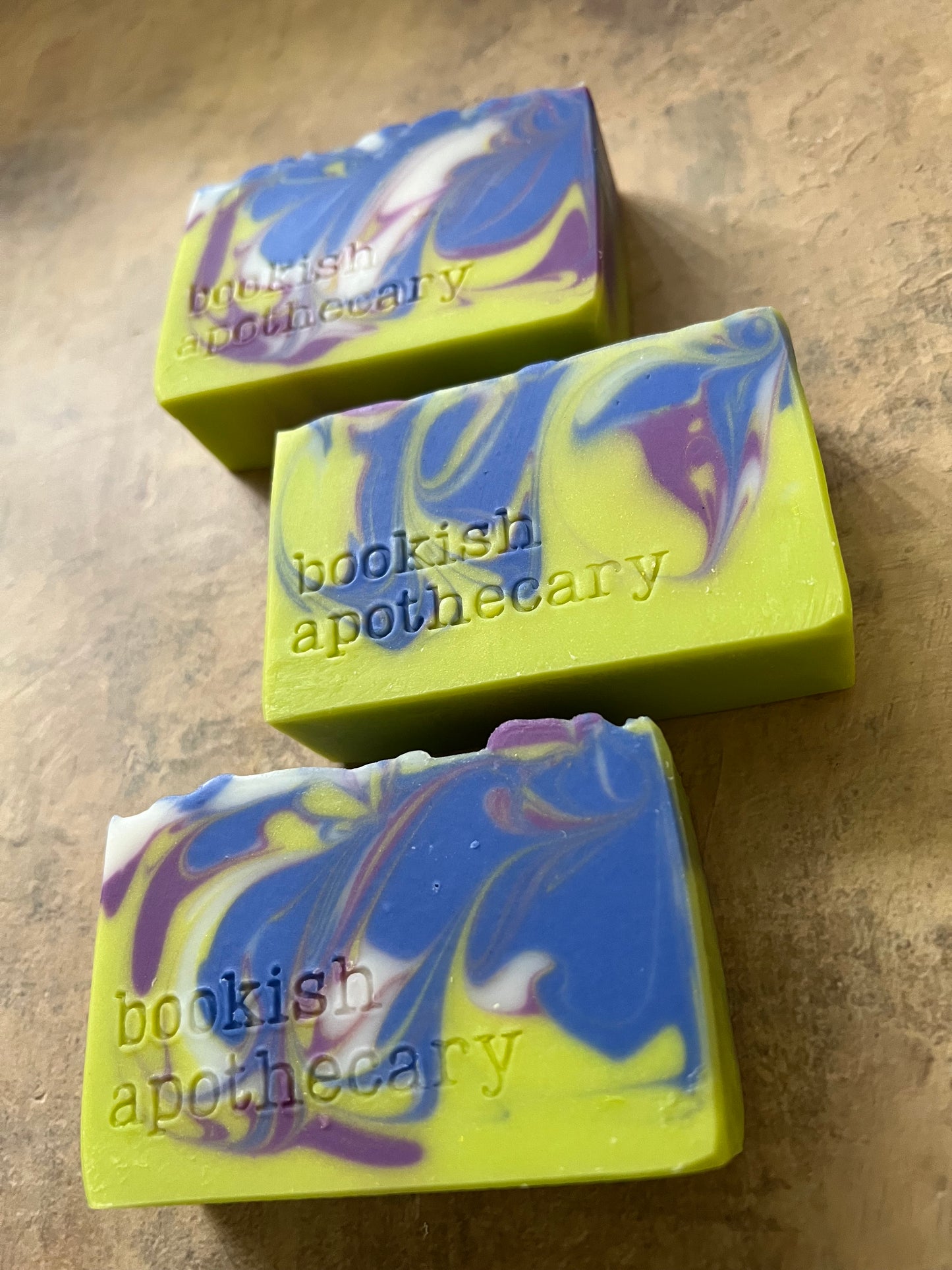 Three rectangular artisanal soaps with a lime green base and blue, purple, and white marbled swirls, displayed on a textured beige surface. The soaps are branded with 'bookish apothecary'.