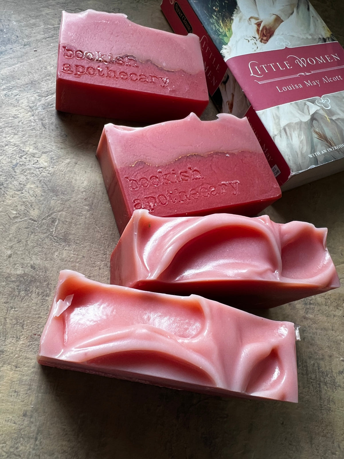 Four pink handmade soap bars with embossed 'bookish apothecary' branding arranged on a textured brown surface, alongside a book titled 'Little Women' by Laura May Abbott.