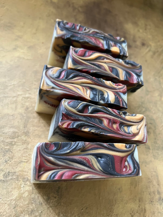 Five rectangular bars of handmade soap with a marbled swirl pattern of black, red, gold, and white, arranged on a light-colored wooden stand against a textured tan background.