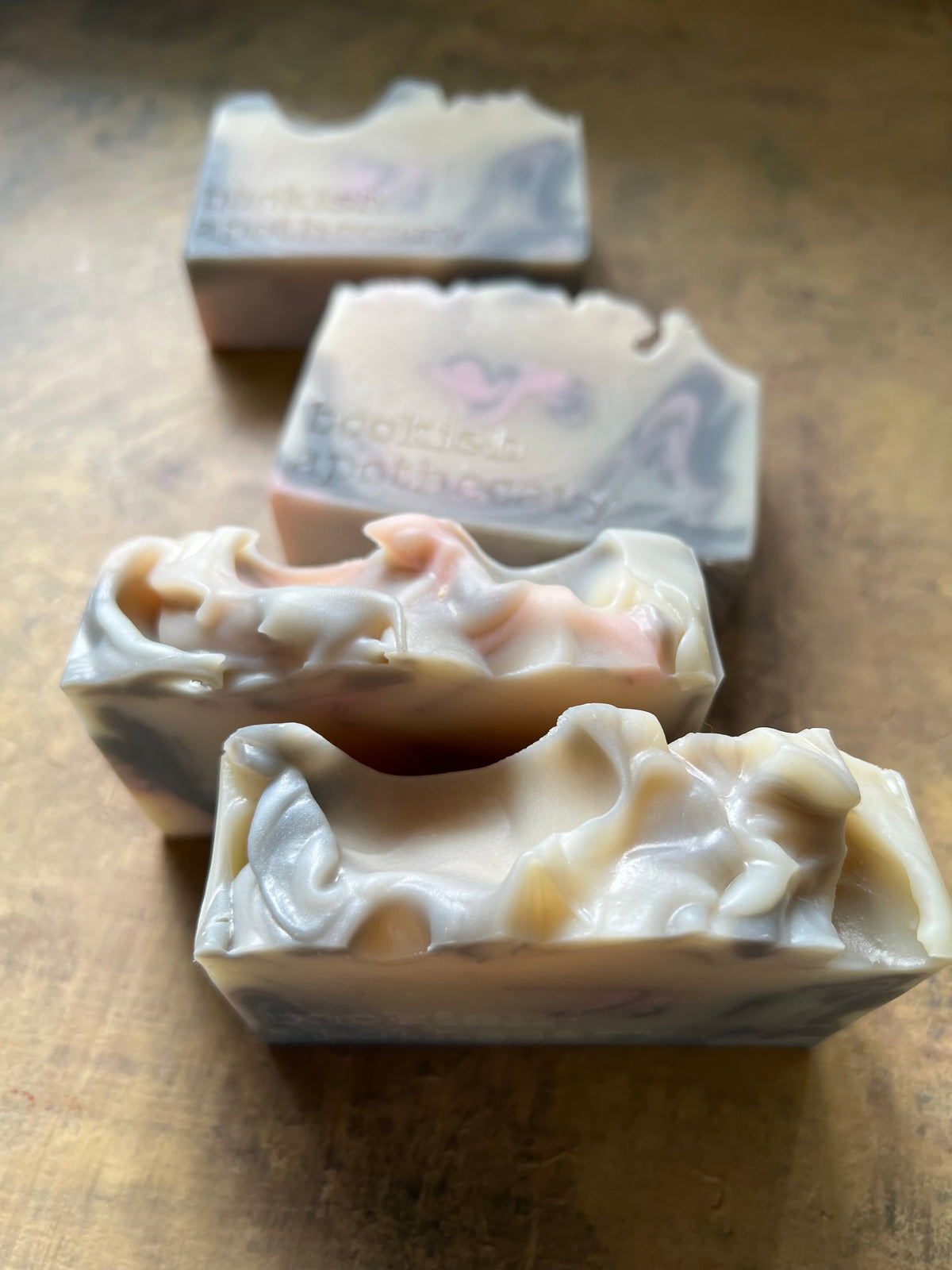 Four handmade soap bars with a marbled cream, pink, and blue-gray pattern, arranged diagonally on a rustic wooden surface. The front soap bar is cut to show the textured, swirled interior.