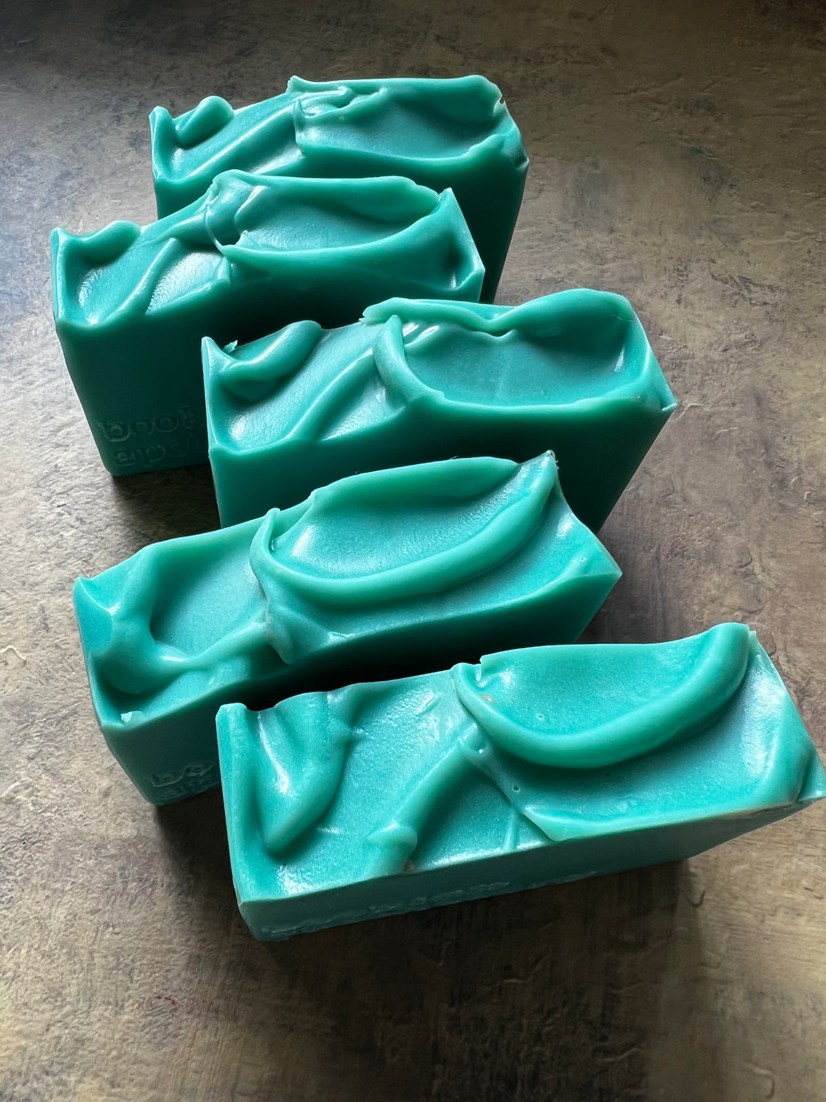 Five handmade  green soap bars with leaf-like embossed patterns, arranged diagonally on a textured brown surface.