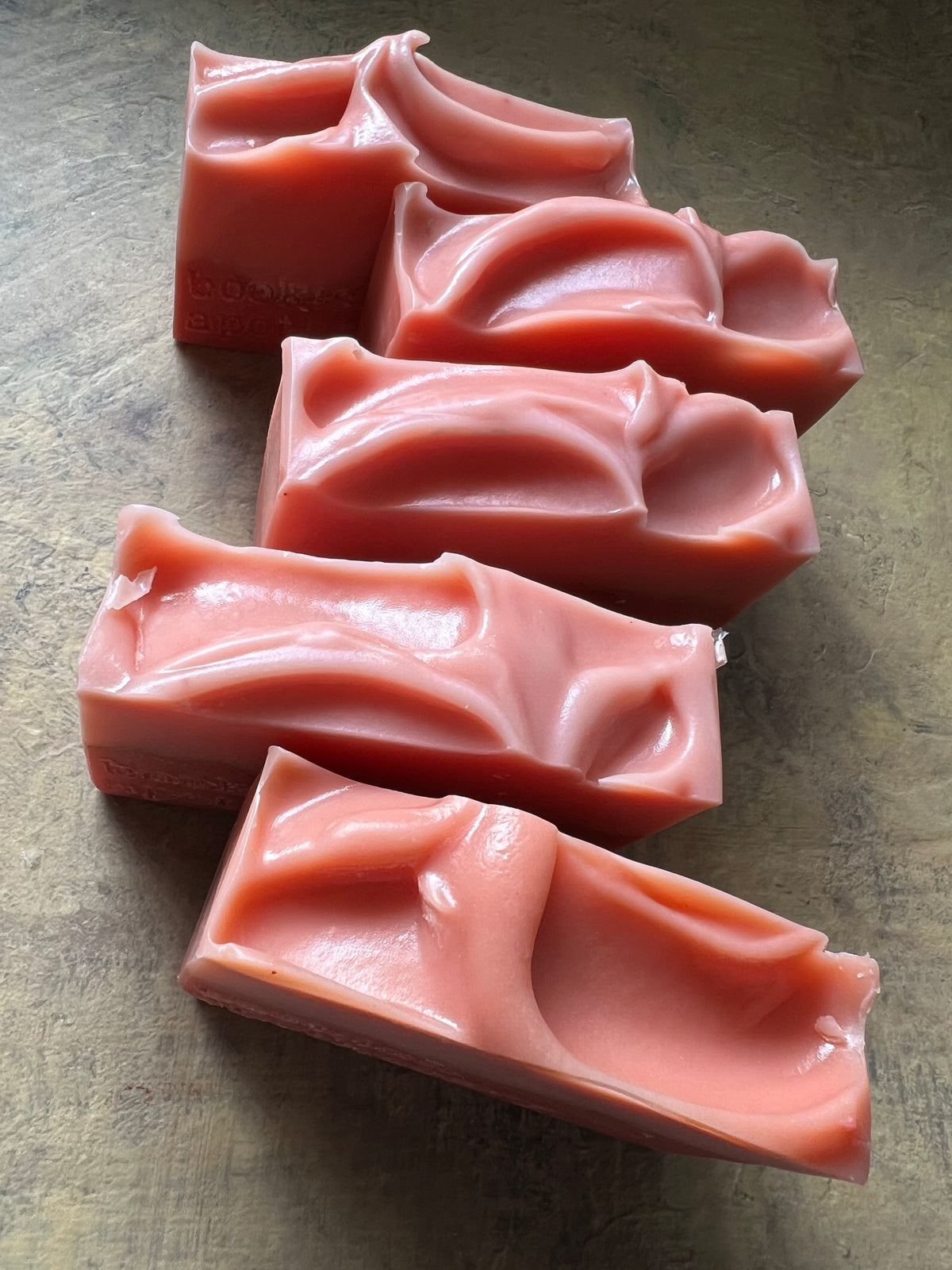 Five handmade soap bars with a wavy, organic texture in a dusty rose pink color, arranged diagonally on a textured gray surface.