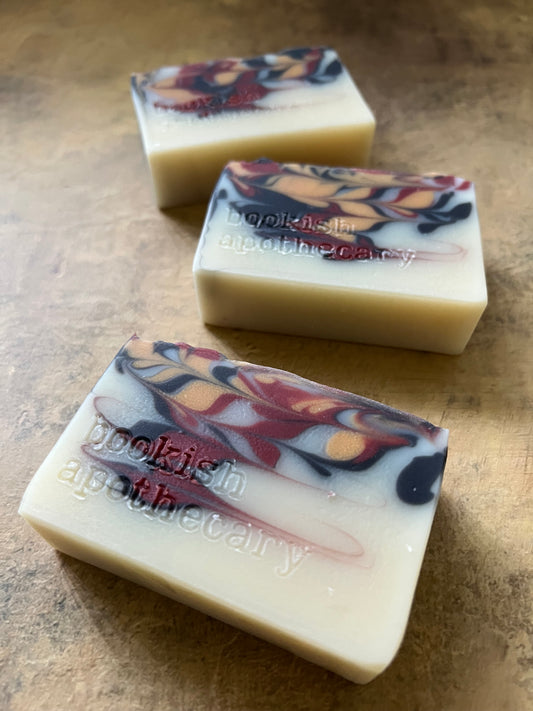Three handmade rectangular bars of soap with cream-colored bases and marbled swirls of red, black, and orange, resting on a textured brown surface. The soap features the brand name 'Bookish Apothecary' embossed vertically.