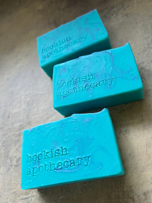 Three turquoise blue soap bars with purple marbling and the brand name 'bookish apothecary' stamped on them, arranged on a textured beige surface.