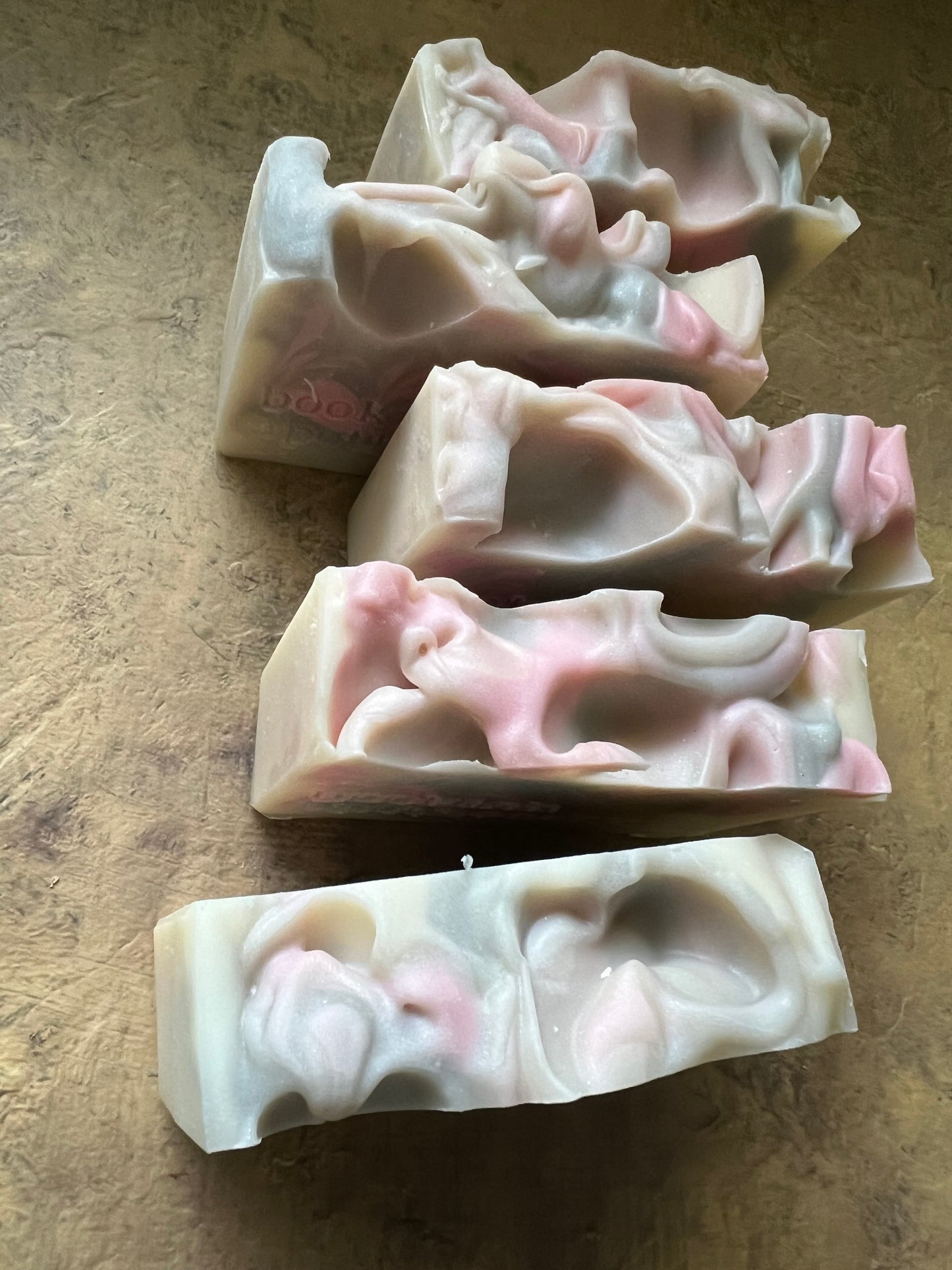 Five handmade soap bars with organic, textured tops  in shades of pink, cream, and gray, arranged on a textured brown surface.