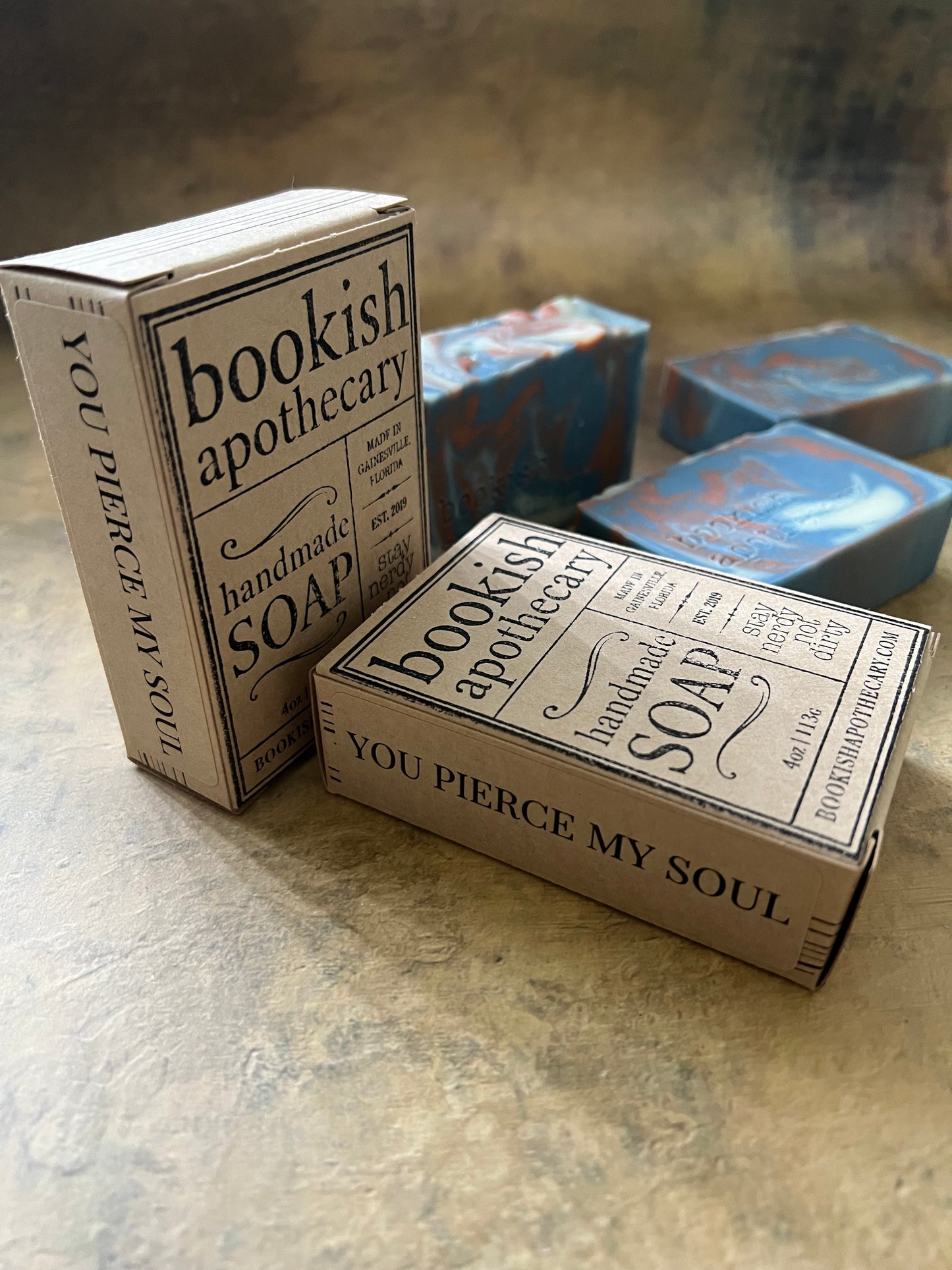 Three vintage-style soap bars wrapped in illustrated paper sleeves labeled 'Bookish Apothecary' and 'You Pierce My Soul,' arranged on a textured beige surface.