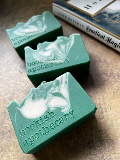 Three handmade green soap bars with white swirls are arranged diagonally on a textured brown surface next to a book.