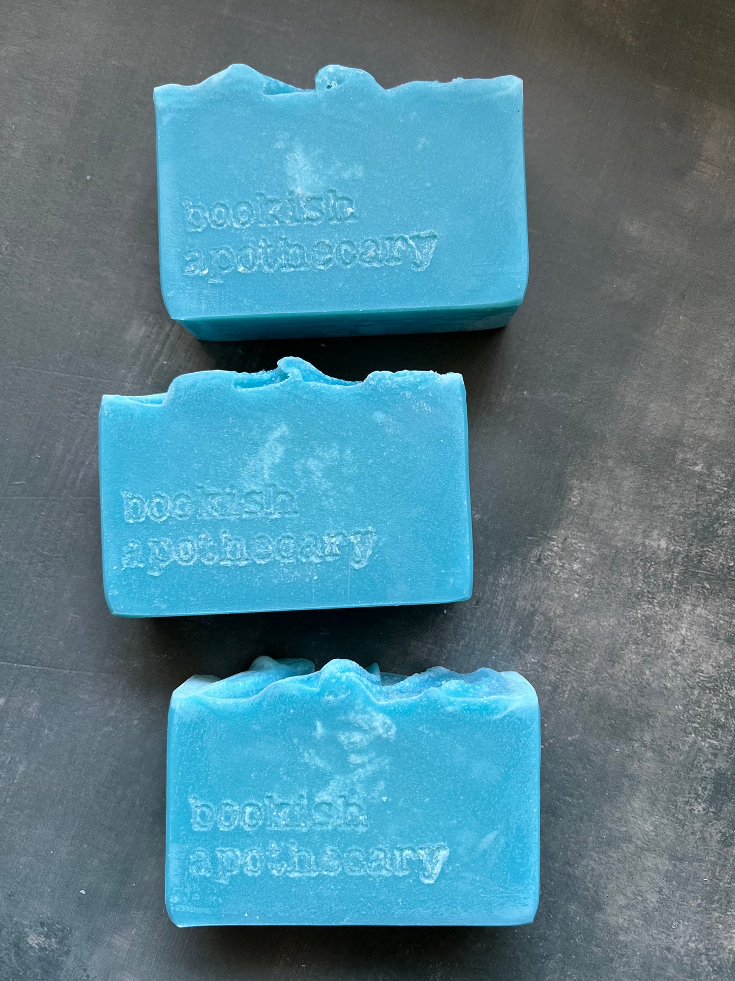 Three blue soap bars with 'Bookish apothecary' branding on a dark surface