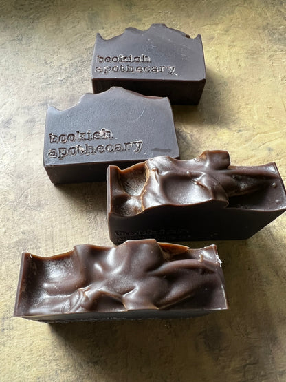 Four dark brown handmade soap bars with textured, organic shapes and embossed 'bookish apothecary' branding, arranged on a textured gold surface.