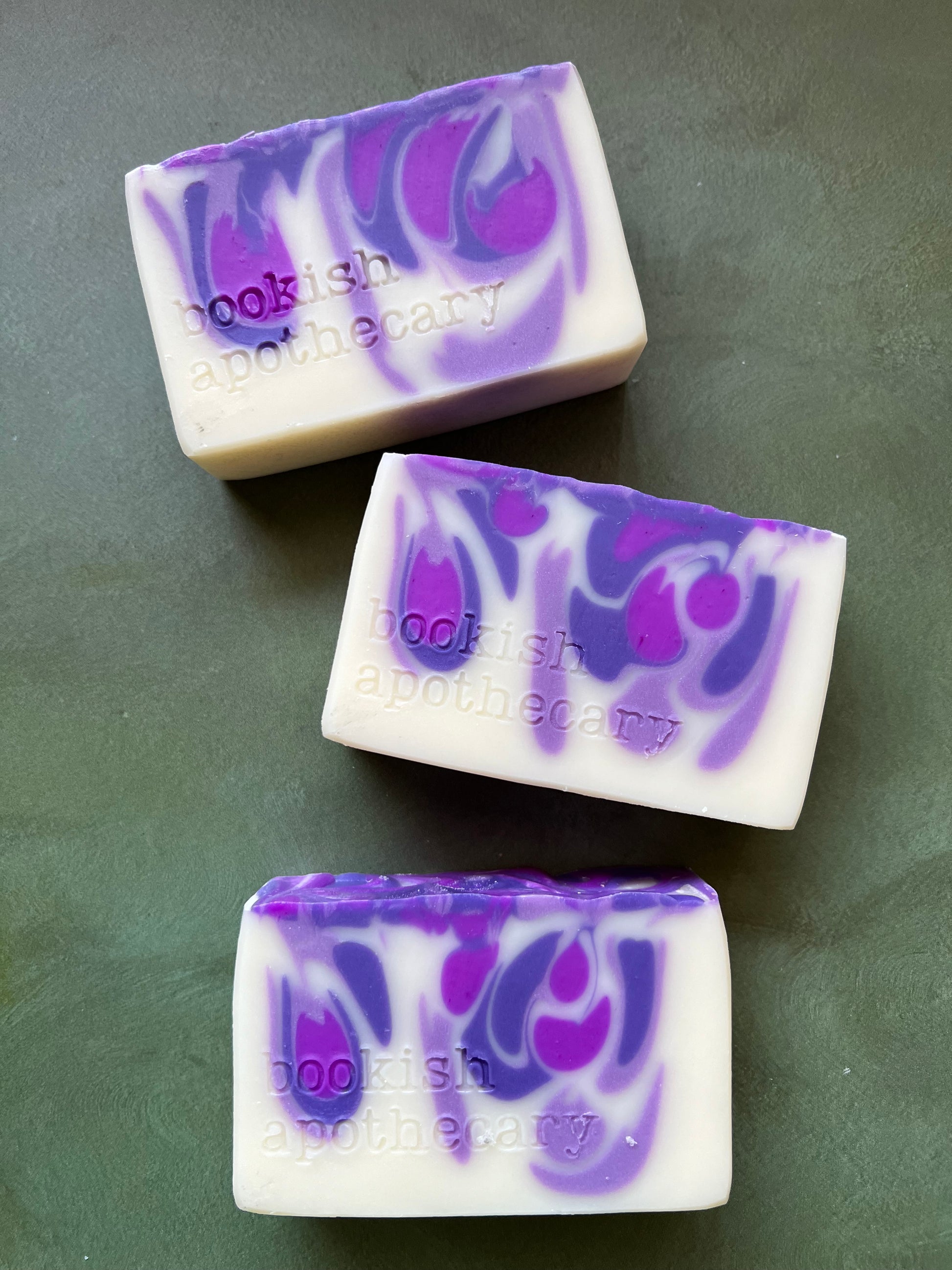 Three bars of white soap with purple swirls stamped with bookish apothecary branding.