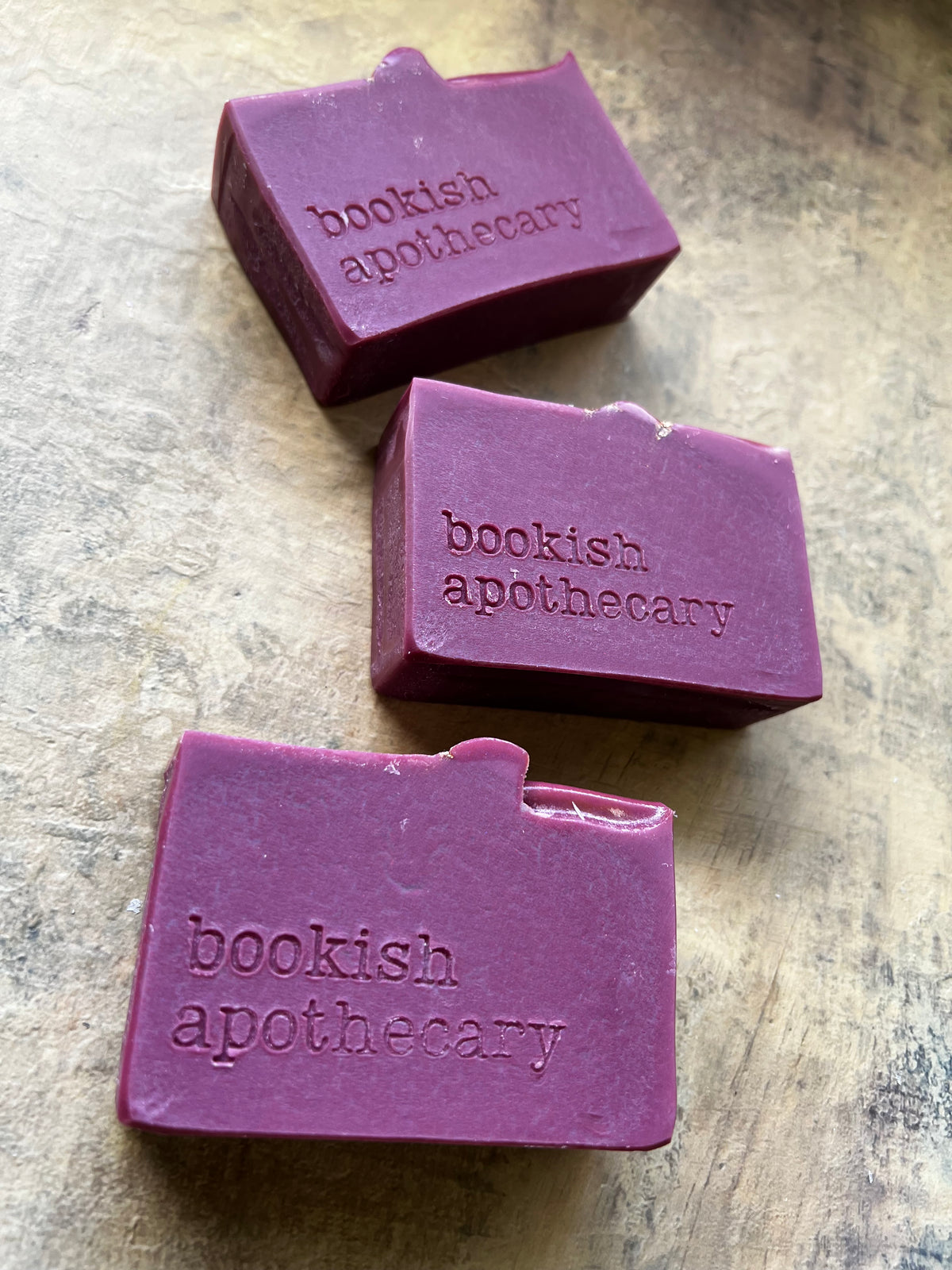 Three rectangular purple soap bars with a small tab on the top edge, each embossed with the brand name 'bookish apothecary', arranged diagonally on a textured beige stone surface.