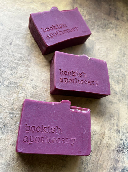 Three rectangular purple soap bars with a small tab on the top edge, each embossed with the brand name 'bookish apothecary', arranged diagonally on a textured beige stone surface.