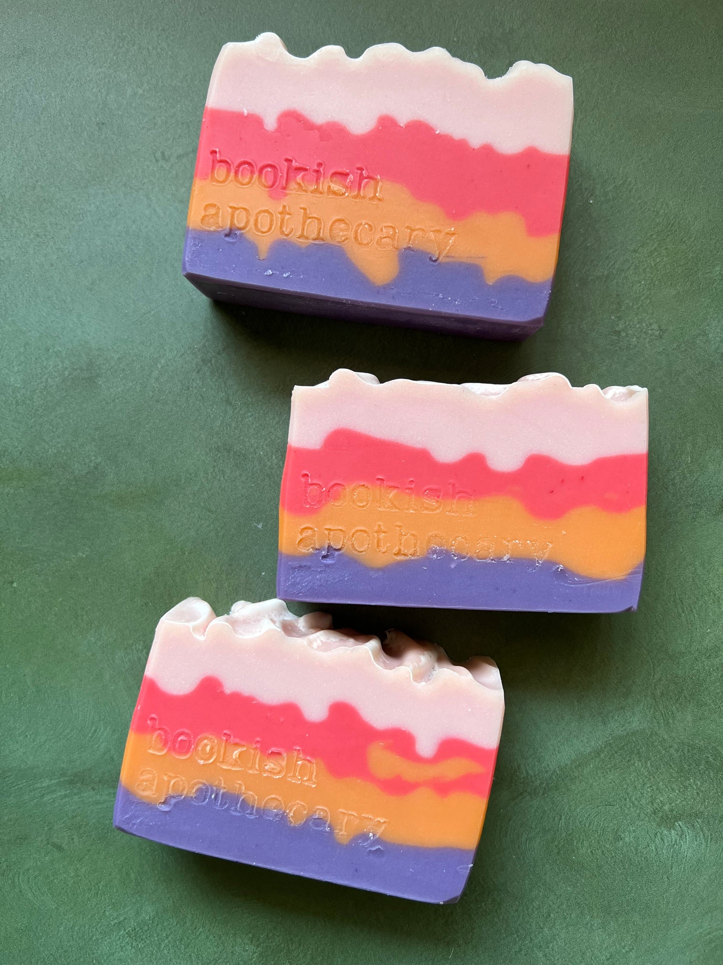 Three bars of soap with layers of pink, red, orange and purple stamped with bookish apothecary branding.