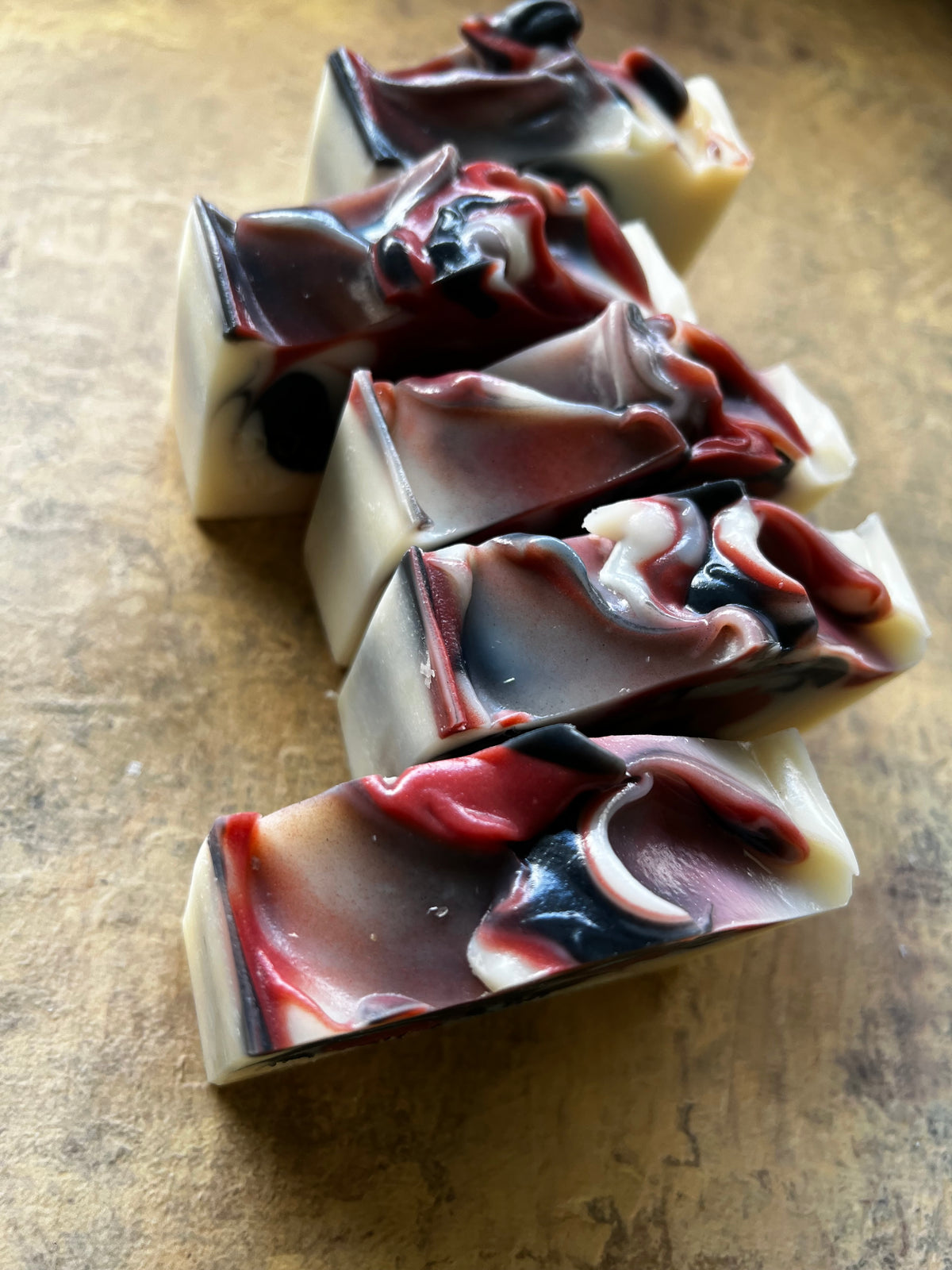 Five rectangular bars of marbled artisanal soap arranged diagonally on a textured brown surface. The soap features swirls of cream, deep red, black.