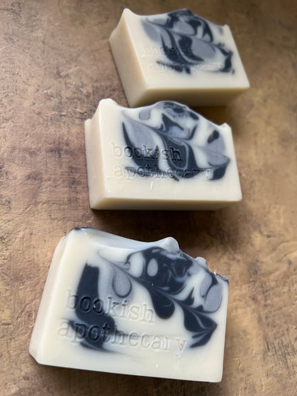 Three rectangular bars of soap with a marbled black and gray pattern on an off-white base, arranged diagonally on a textured brown surface. The bars are stamped with the brand name 'bookish apothecary'.