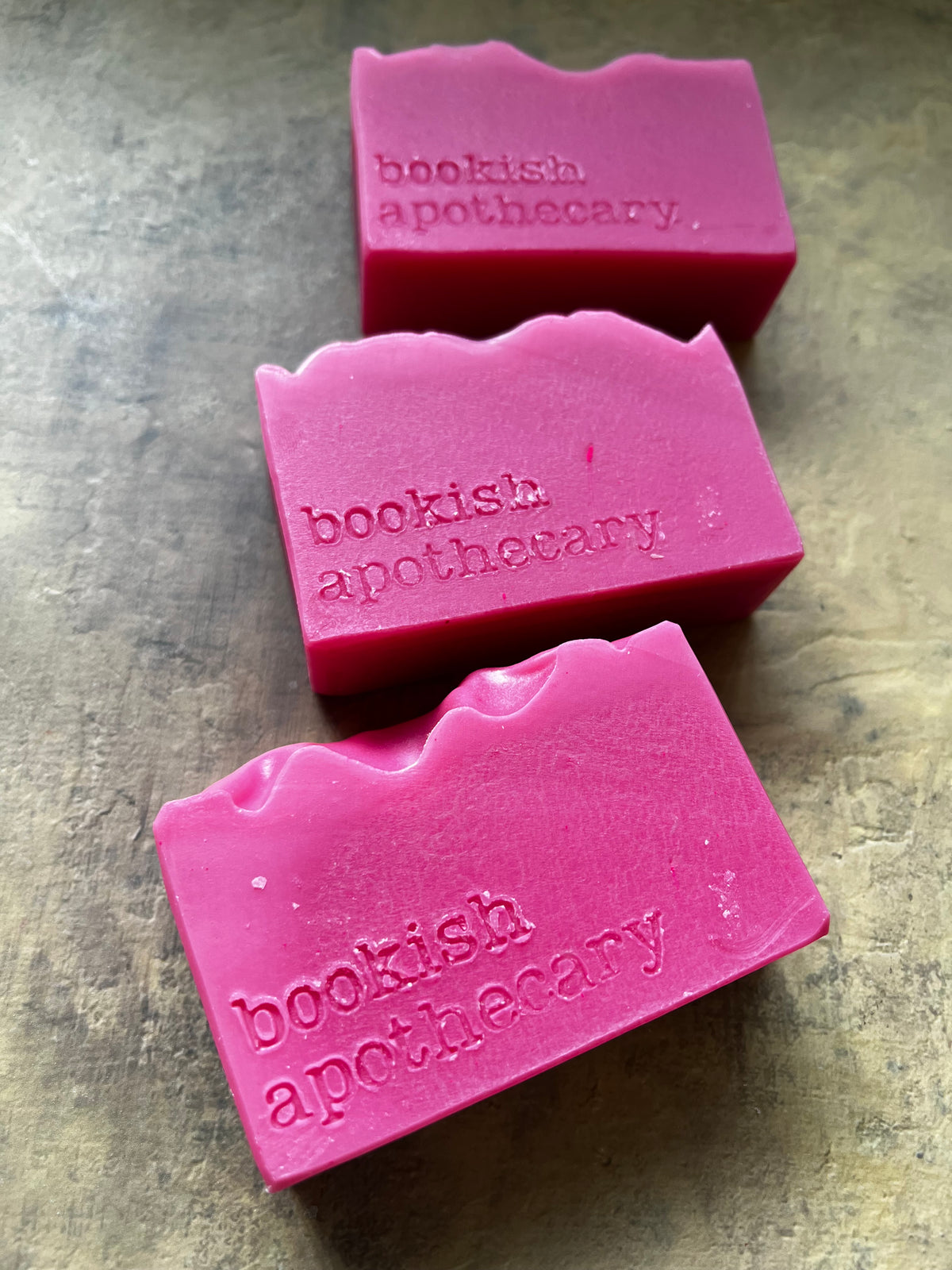 Three rectangular pink soap bars with wavy cut tops, each embossed with the brand name 'bookish apothecary', arranged diagonally on a textured gray surface.