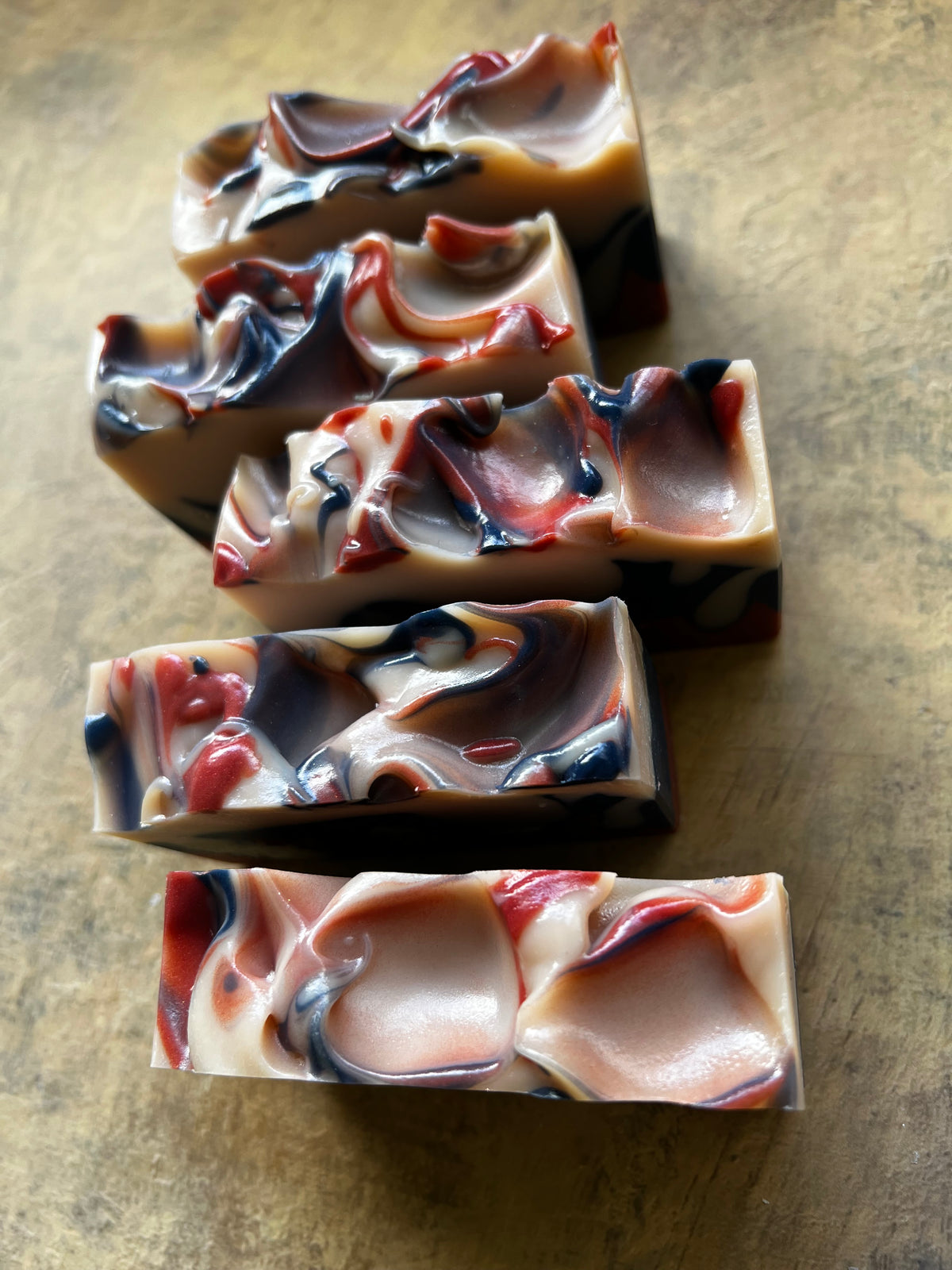 Five handmade marbled soap bars arranged vertically on a textured brown surface. The soaps feature swirls of cream, dusty rose, deep red, and dark navy blue.