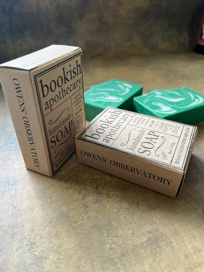 Two  bars of Owens' Observatory handmade bookish apothecary soap in kraft packaging, arranged on a textured gray surface.