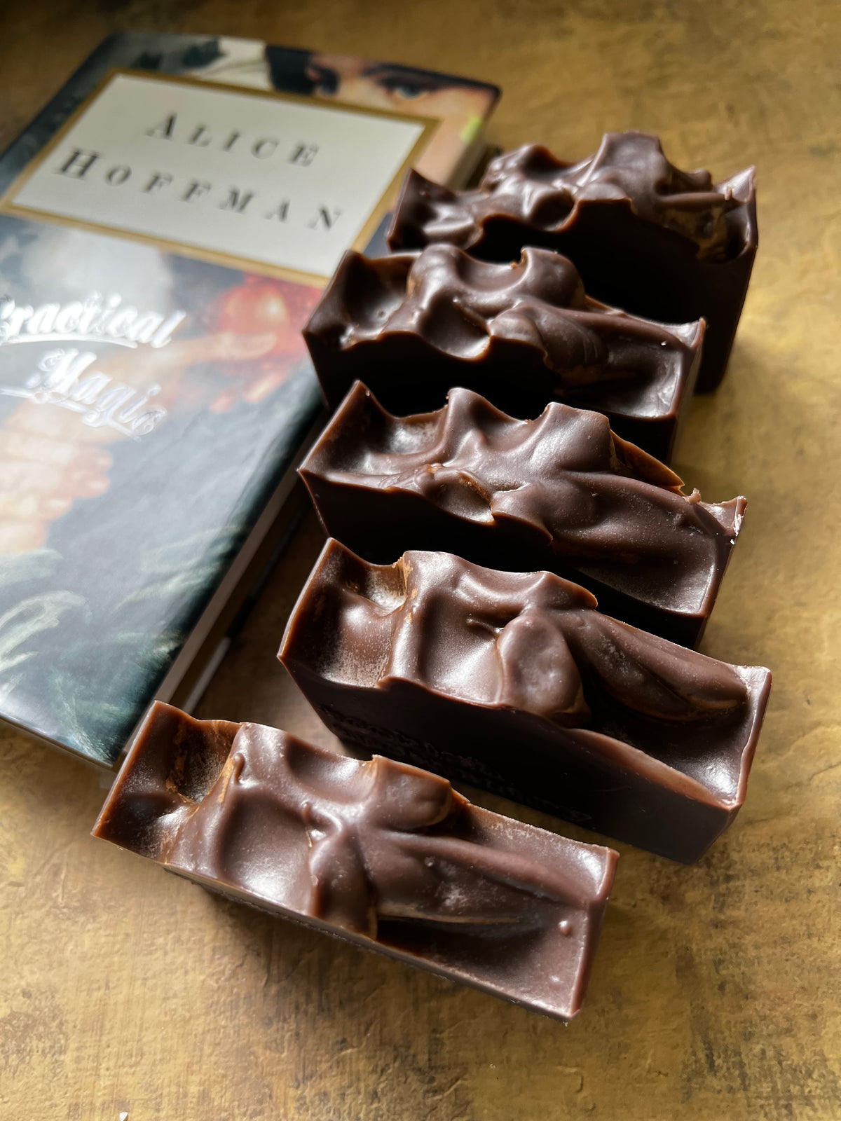 Five dark chocolate bars with a textured, surface are arranged diagonally next to a book titled 'Practical Magic' by Alice Hoffman on a rustic, metallic gold surface.