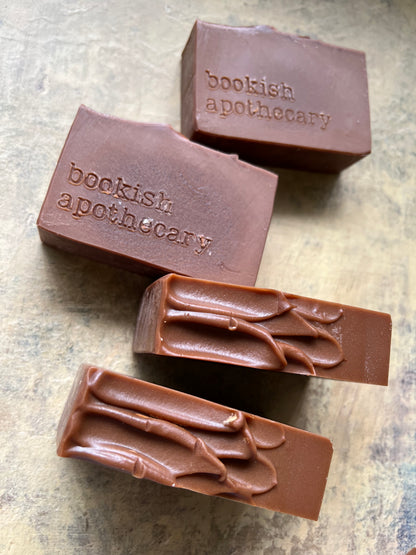 Four rectangular bars of brown soap with the brand name 'bookish apothecary' on two bars, arranged on a textured gray surface.