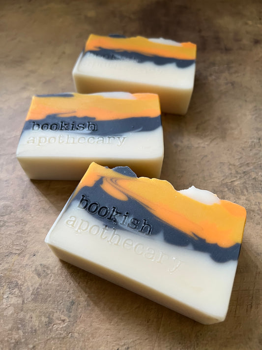 Three handmade soap bars with a cream base and orange, navy, and gray marbled swirls, resting on a textured brown surface. The brand name 'hookish apothecary' is stamped on the soaps.