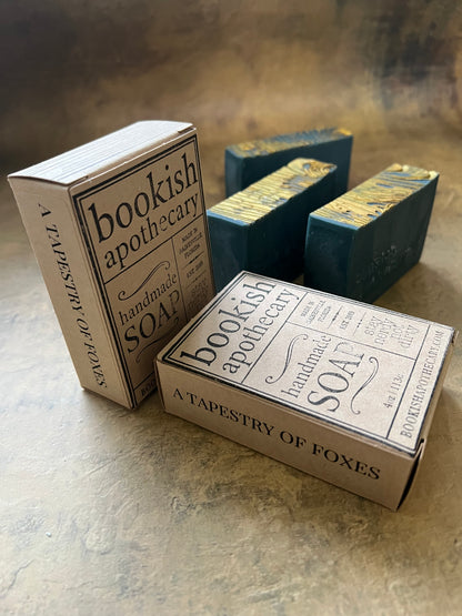 Three handmade soap bars with decorative packaging boxes arranged on a textured beige surface. The packaging features vintage-style typography reading 'Bookish Apothecary' and 'A Tapestry of Foxes.