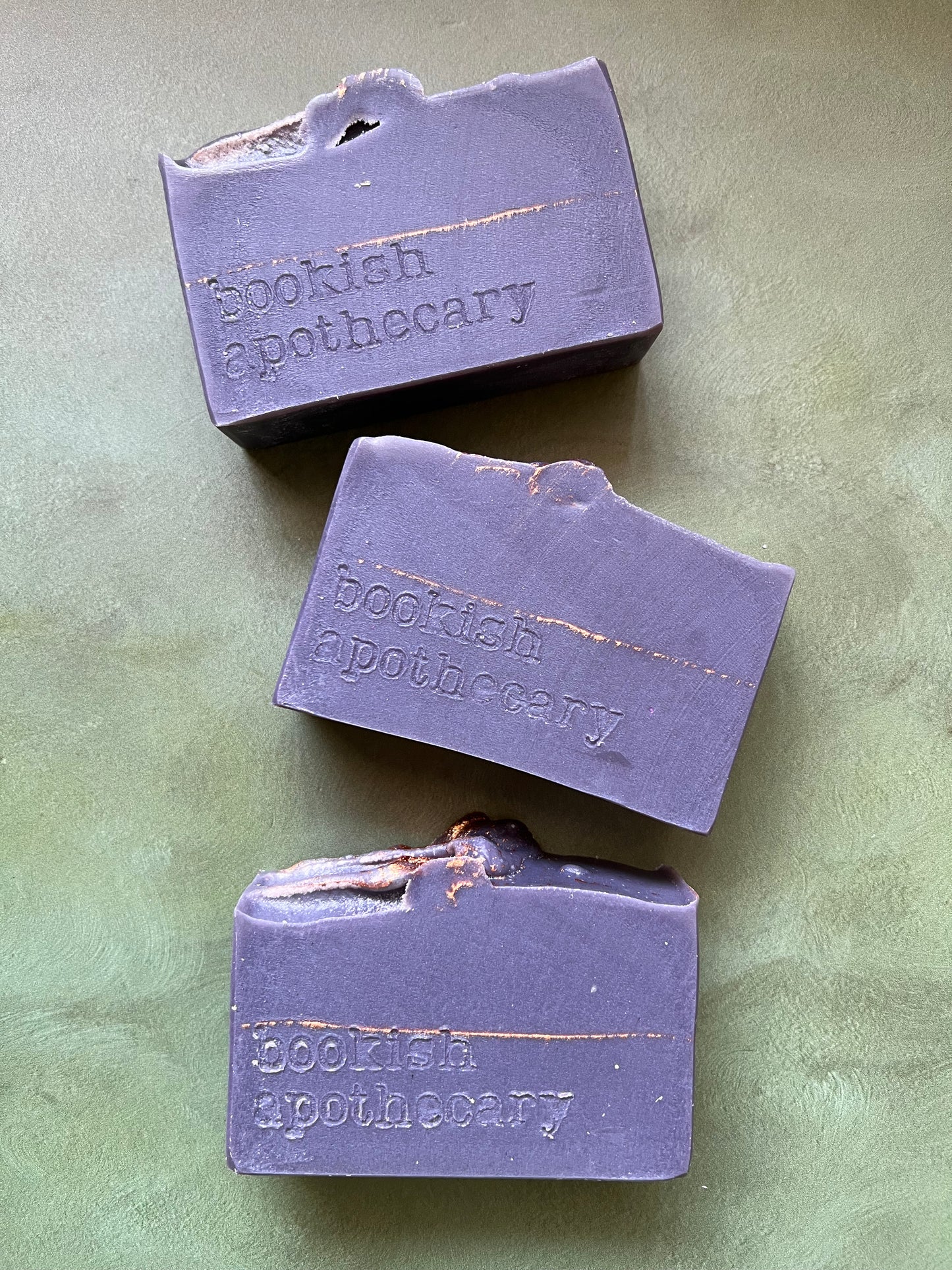 Three bars of dark purple soap stamped with bookish apothecary branding.