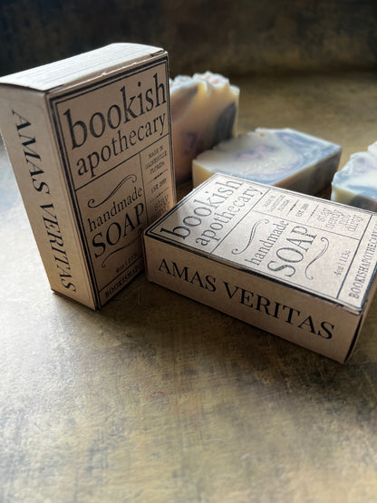 Two handmade soap bars with matching vintage-style paper boxes labeled 'bookish apothecary' and 'AMAS VERITAS' on a textured metallic surface.