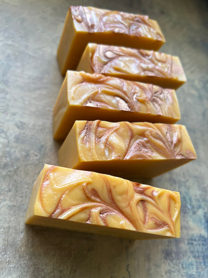 Five rectangular bars of handmade soap with a marbled swirl pattern in warm yellow, orange, and brown, arranged diagonally on a textured gray surface.