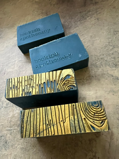 Two dark green rectangular bars of soap standing upright on a textured gold surface and two laying flat with "bookish apothecary" stamped on the side.  