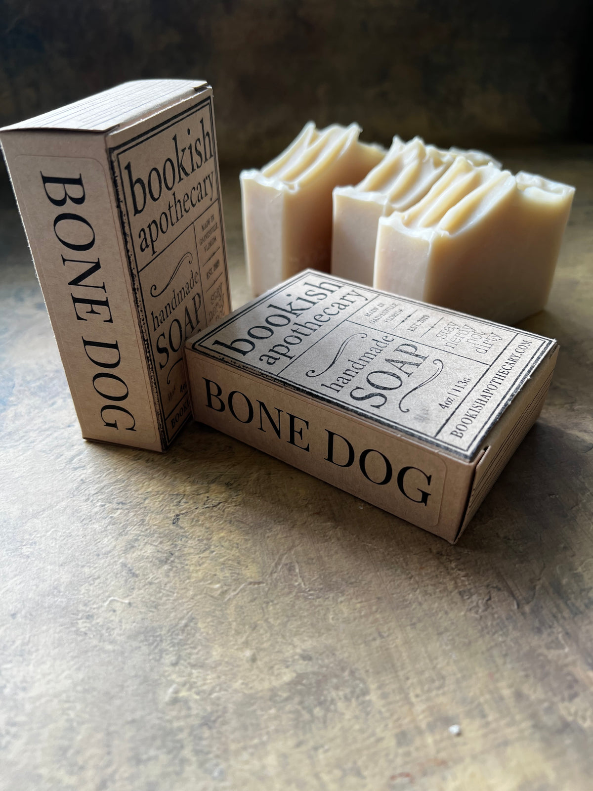 Handmade soap bars in a rustic setting, with two cream-colored bars in the background and two paperboard boxes in the foreground labeled 'BONE DOG' and 'bookish apothecary'.