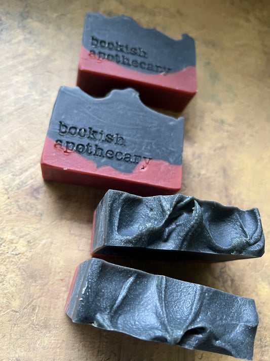 Four handmade soap bars with a dark charcoal gray top and a reddish-brown bottom base, arranged on a textured wooden surface. Two soaps have a wavy cut edge and are stamped with the text 'bookish apothecary'.