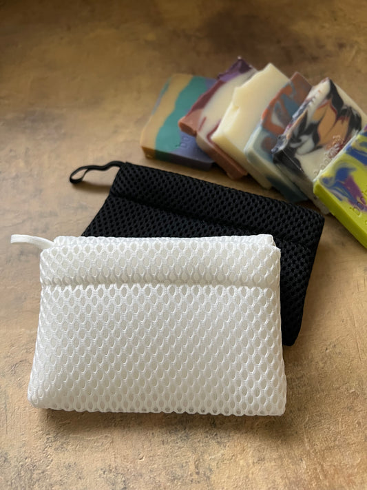 A white textured mesh soap pouch with a black mesh soap mesh envelope, displayed next to a stack of colorful handmade soap bars on a textured beige surface.
