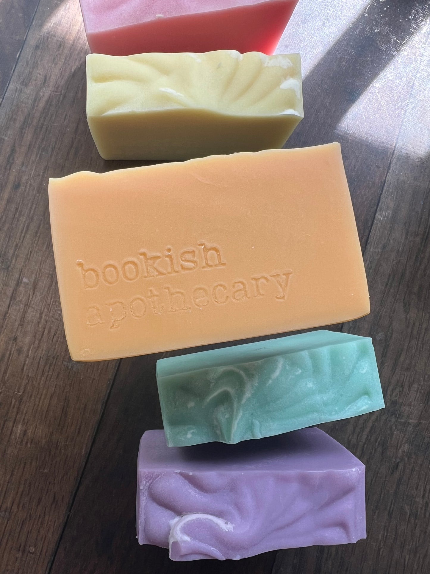 Five bars of soap in different colors on a wooden surface, with 'bookish apothecary' branding.