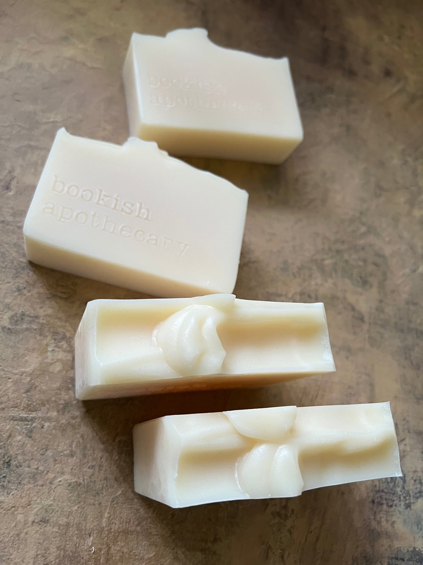 Four off-white handmade soap bars with embossed text and sculpted details, arranged on a textured brown surface.