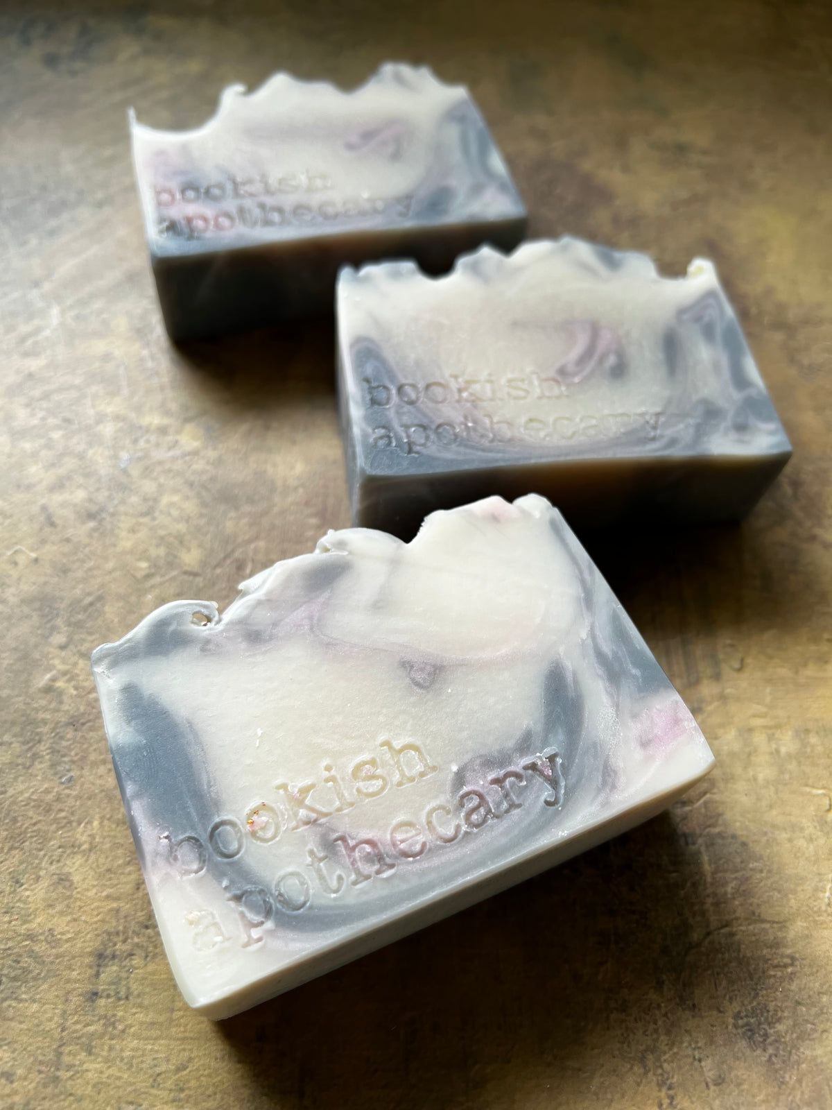 Three handmade soap bars with marbled cream, gray, and pale pink swirls are arranged diagonally on a textured brown surface.