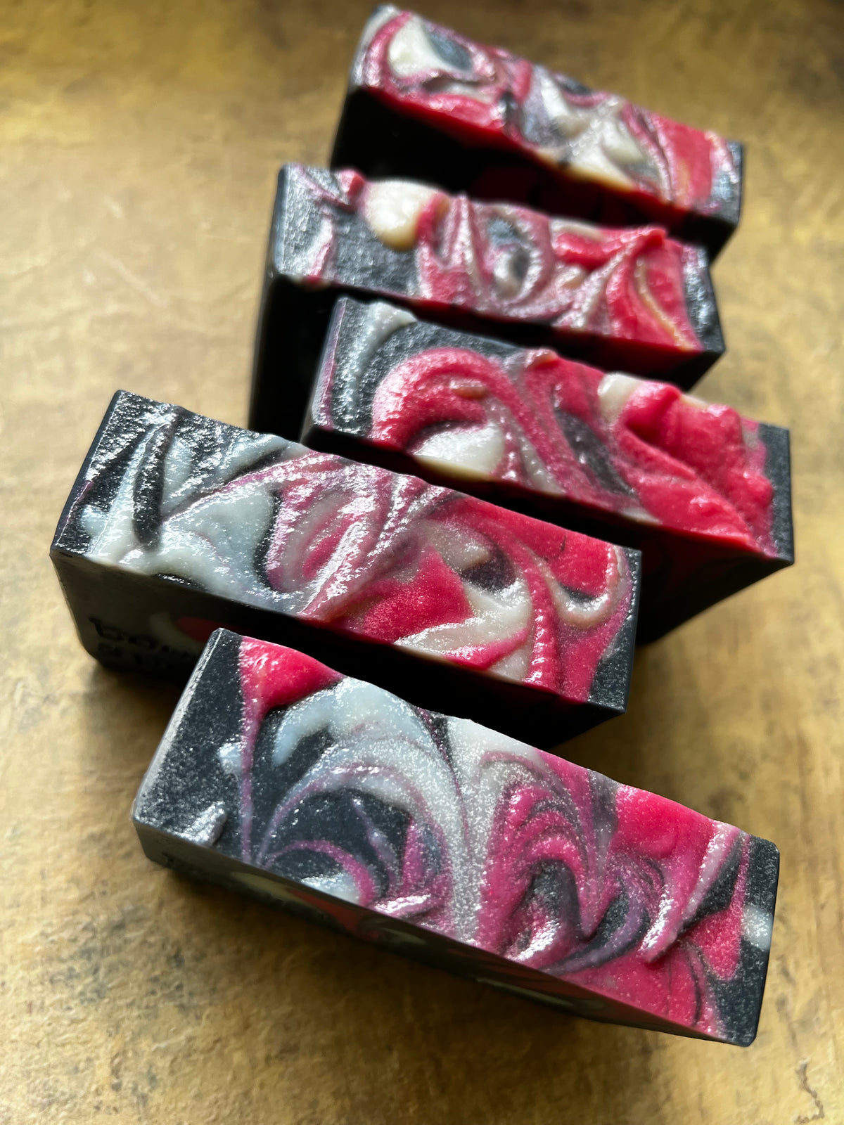 Five rectangular handmade soap bars with a marbled swirl pattern of red, pink, white, and black, arranged diagonally on a textured brown surface.